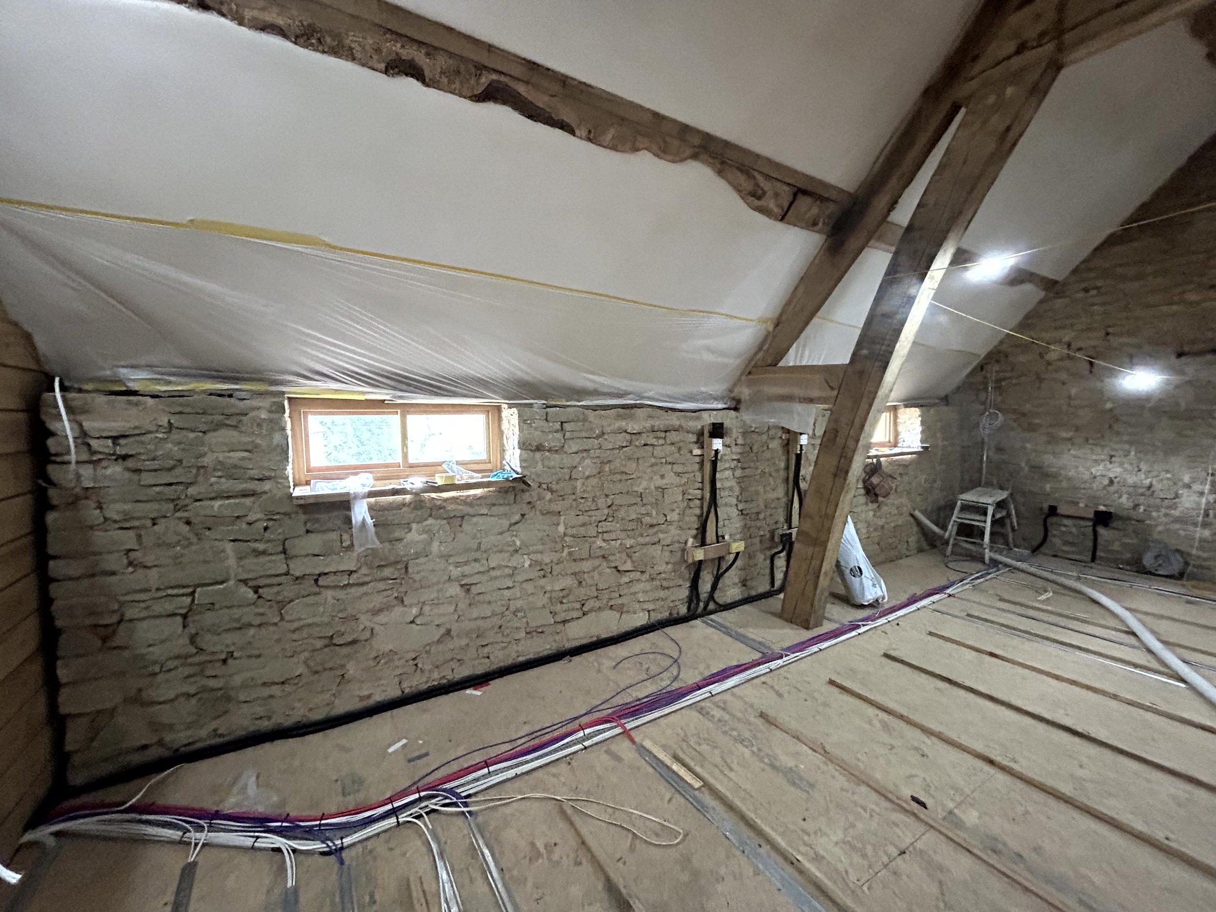 Bare stone gable wall before insulation, plastic sheeting protecting the roof slope, first-fix wiring laid across the subfloor