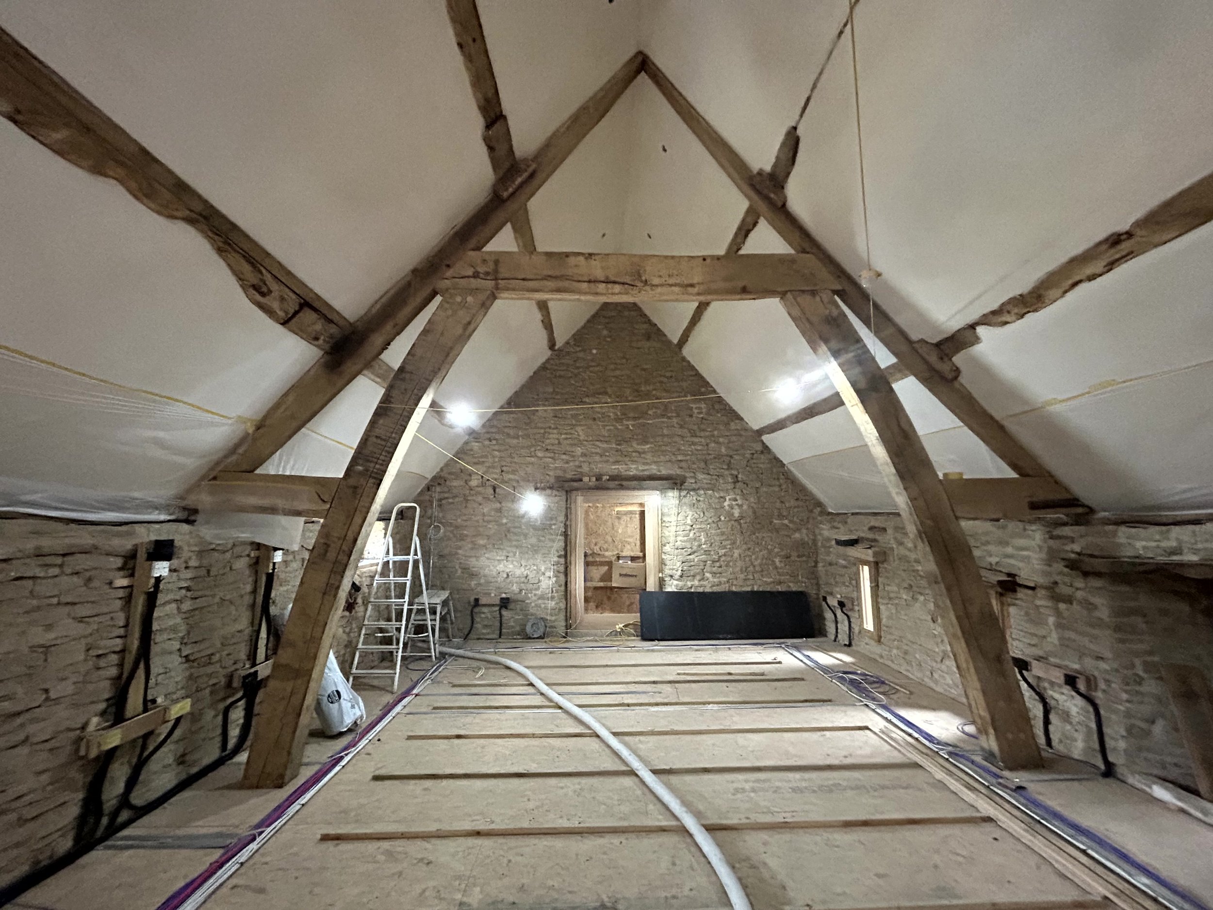 Full view down the barn's roof space showing the oak A-frame trusses and bare stone gable wall before insulation