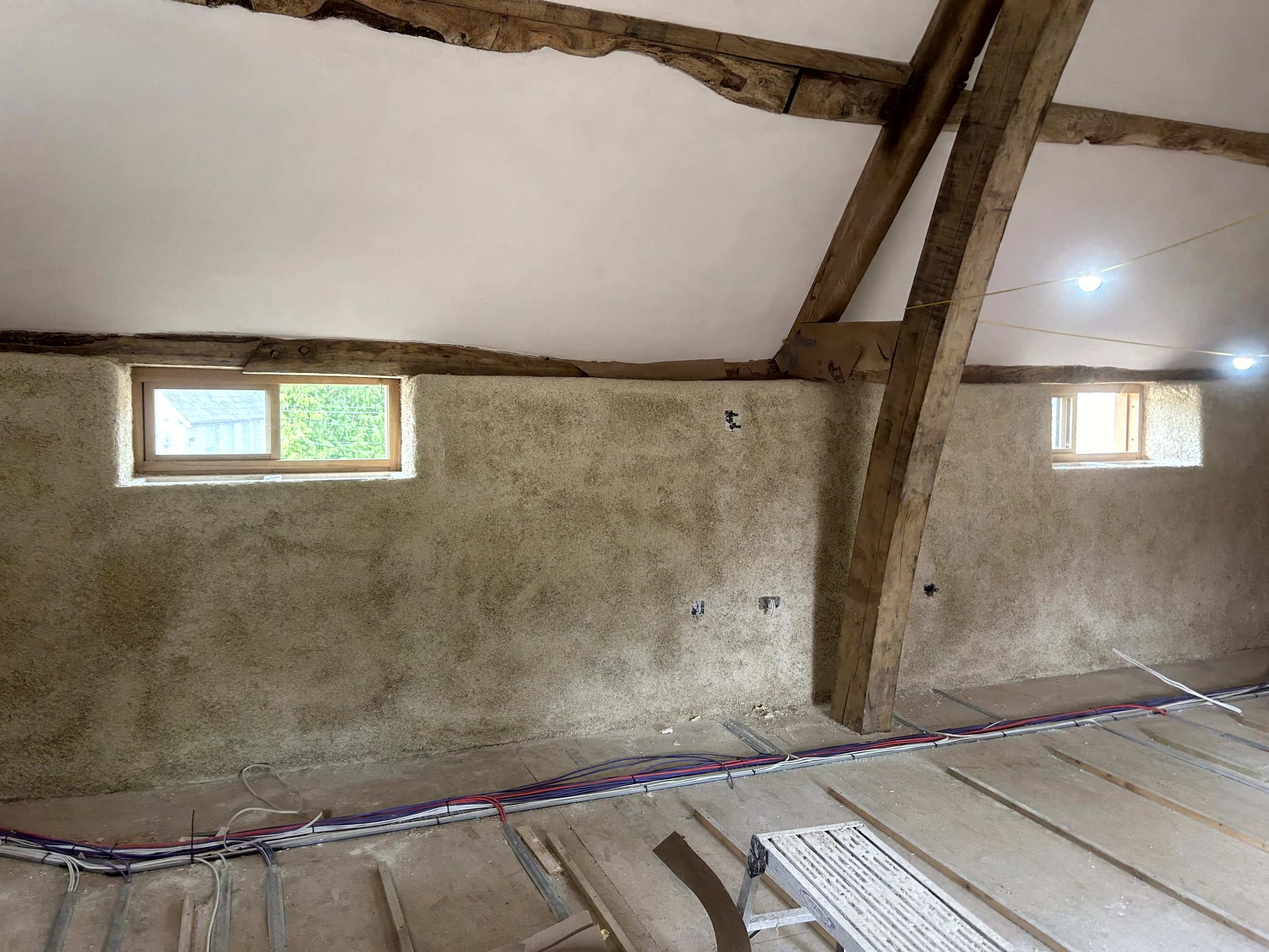 Sprayed hempcrete wall with two windows of different sizes, exposed roof beams above, cabling still laid across the subfloor